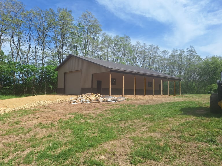 Brown metal barn with covered porch and wooden posts set in a grassy field with trees in the background