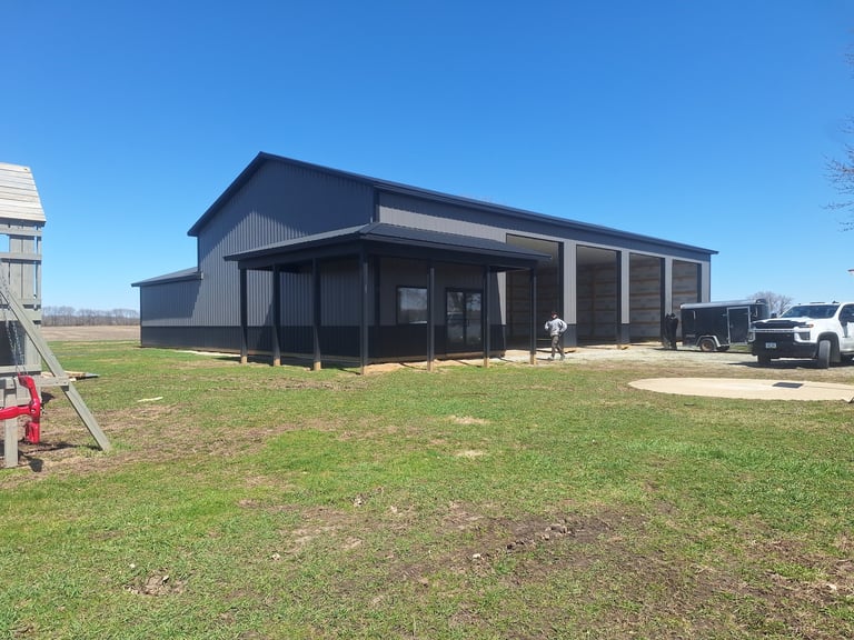 Modern dark blue metal-frame agricultural building with covered porch on grassy rural property with vehicles and structures nearby